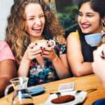 Four people are sitting at a wooden table, laughing and enjoying coffee together. There is a plate with a pastry and a pitcher of coffee on the table.
