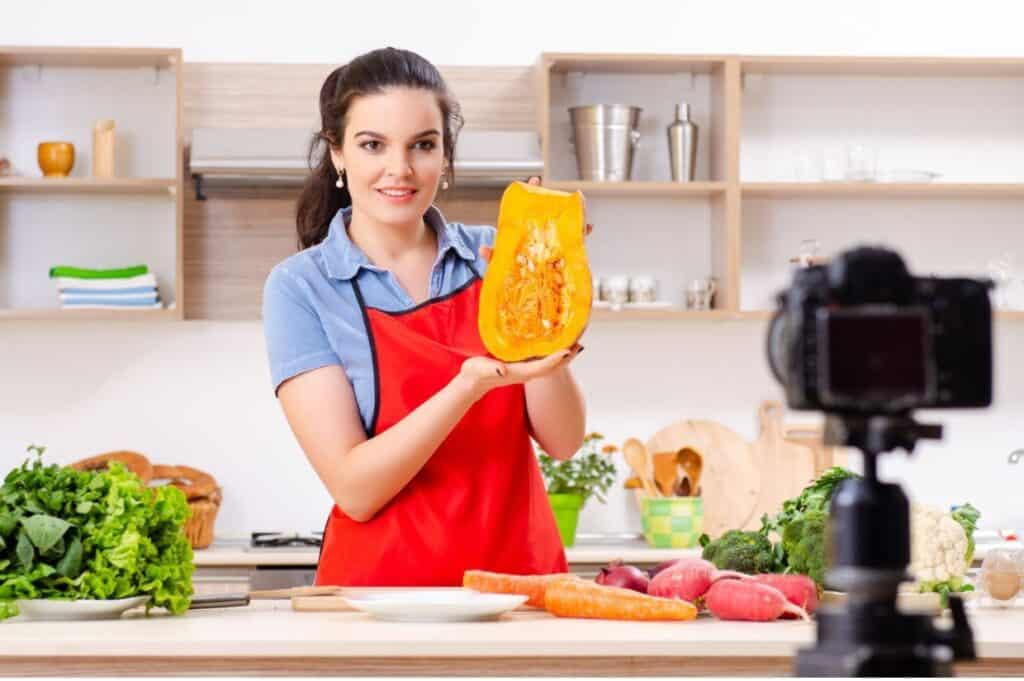 A woman in a red apron holds a halved pumpkin in a kitchen, standing in front of a camera. The kitchen counter in front of her has vegetables like carrots and greens. Shelves with kitchenware are in the background.