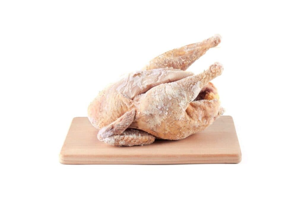 Frozen whole chicken placed on a wooden cutting board against a plain white background.