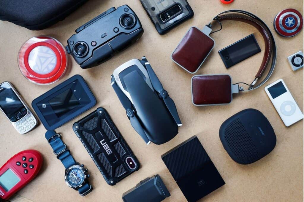 A collection of various electronic gadgets and accessories arranged neatly on a surface, including a drone, smartwatch, headphones, portable speaker, camera, smartphone, and other tech items.