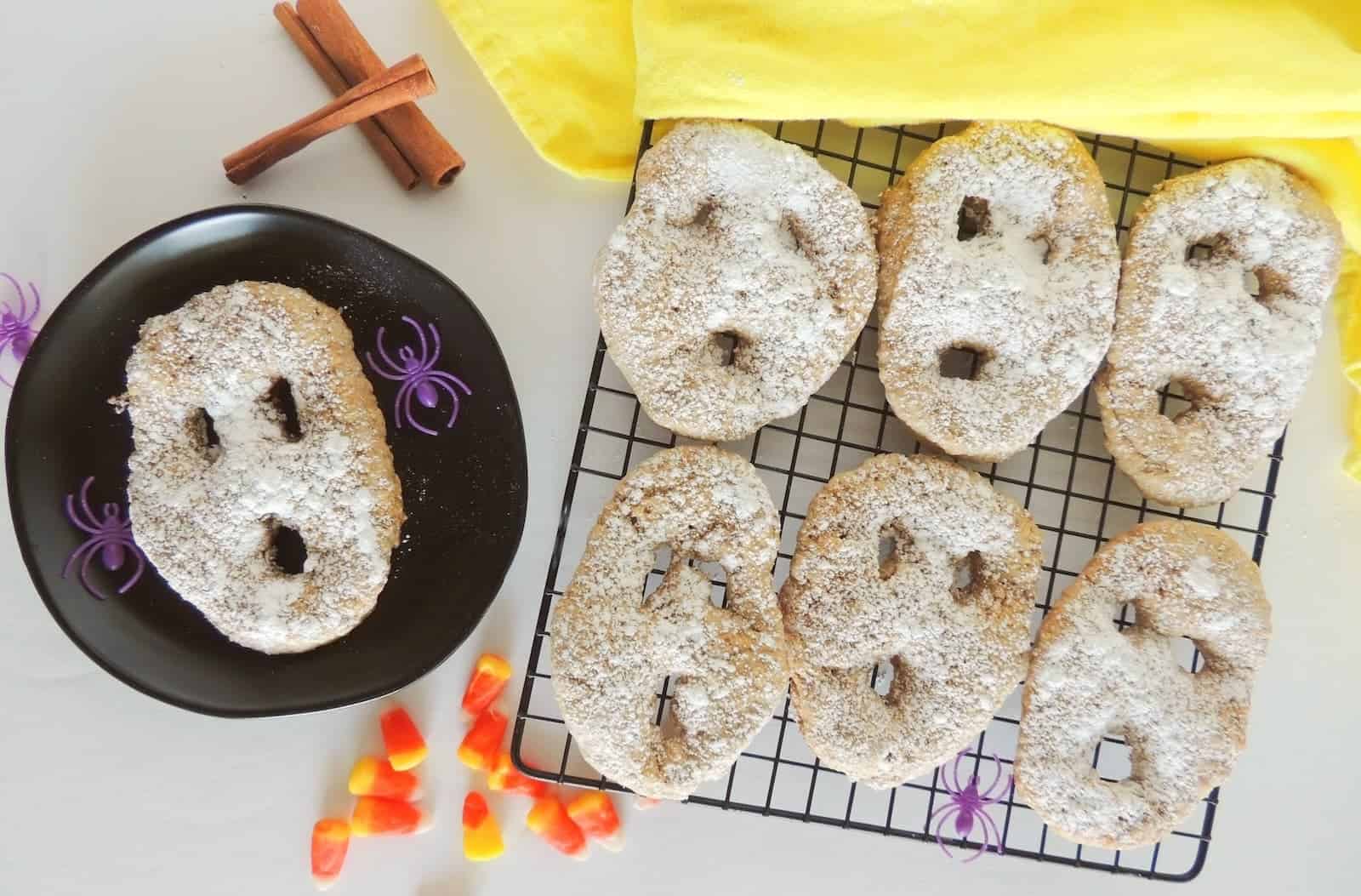 Halloween-themed scones shaped like ghosts, arranged on a cooling rack and a black plate. Cinnamon sticks, yellow cloth, and candy corn are nearby. The scones are dusted with powdered sugar.