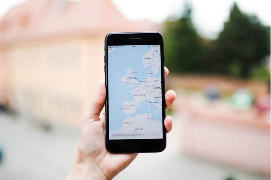 A hand holding a smartphone displays a map of Europe with Mediterranean and Northern Europe visible, blurred buildings and greenery in the background.