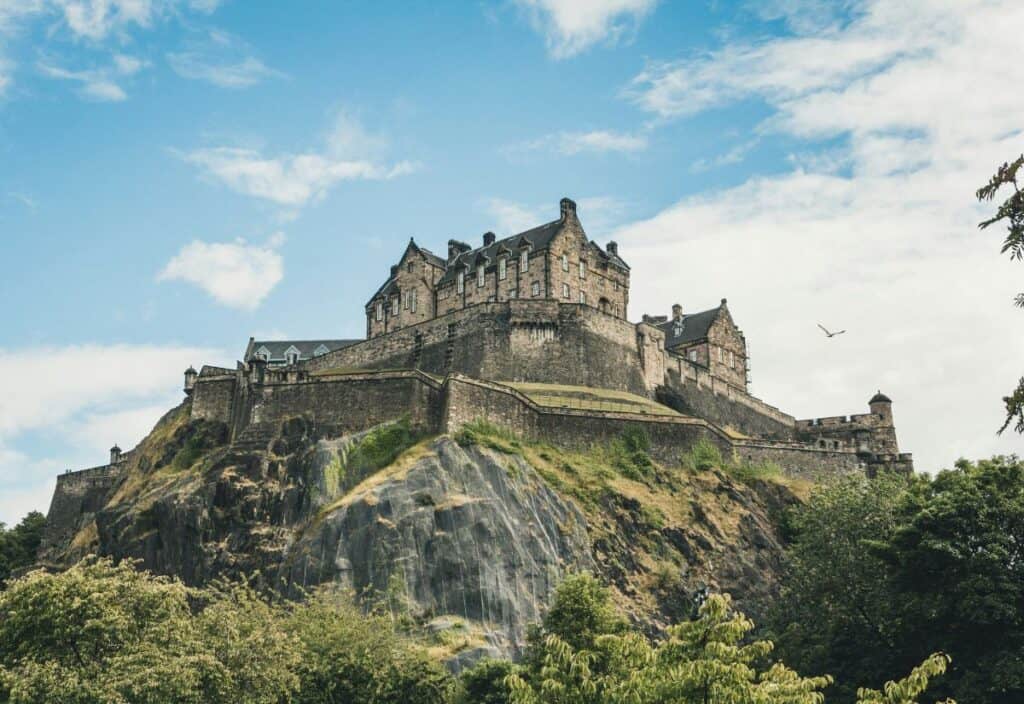 A historic stone castle sits atop a rocky hill with a cloudy sky and a bird flying nearby. Trees and greenery surround the base.