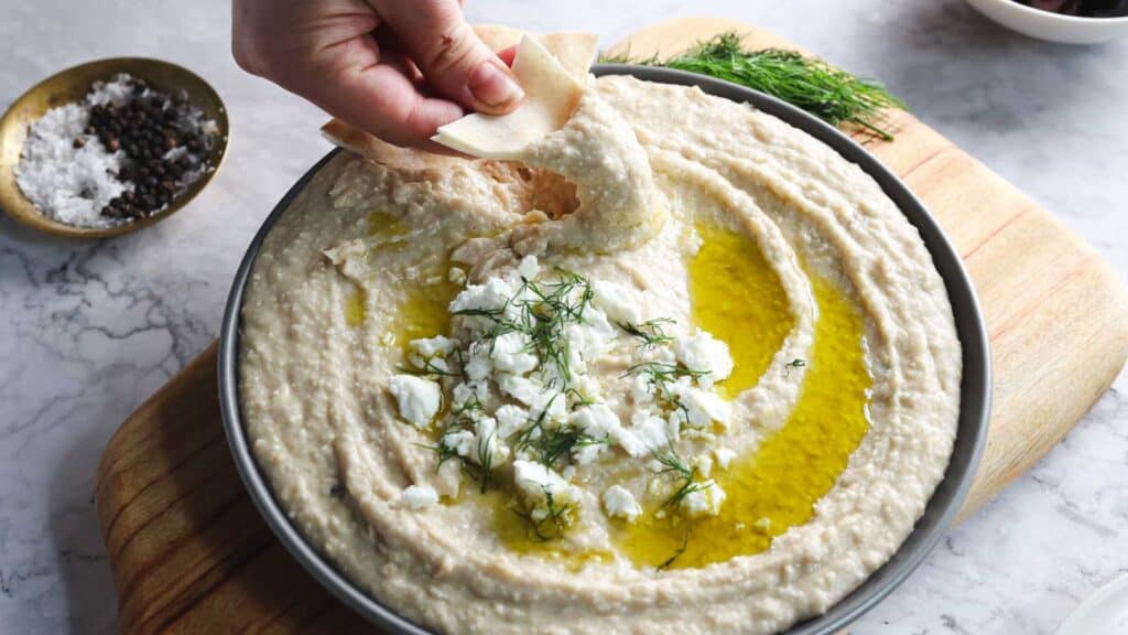 A hand dips pita bread into a bowl of hummus topped with olive oil, crumbled feta cheese, and fresh herbs. Ground pepper and sea salt are in a small bowl on the side.