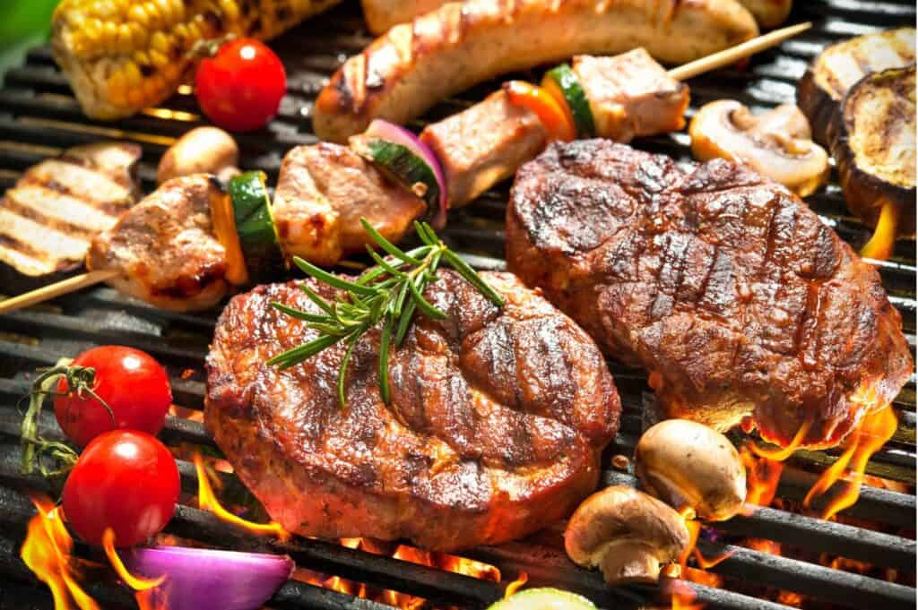 A grill loaded with steaks, sausages, vegetable skewers, cherry tomatoes, corn, mushrooms, and eggplant slices.