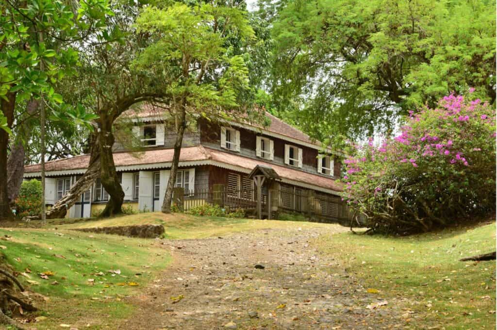 A rustic two-story house with a brown roof set amidst Martinique's lush landscapes and blooming flowers, featuring a pathway leading up to its entrance.