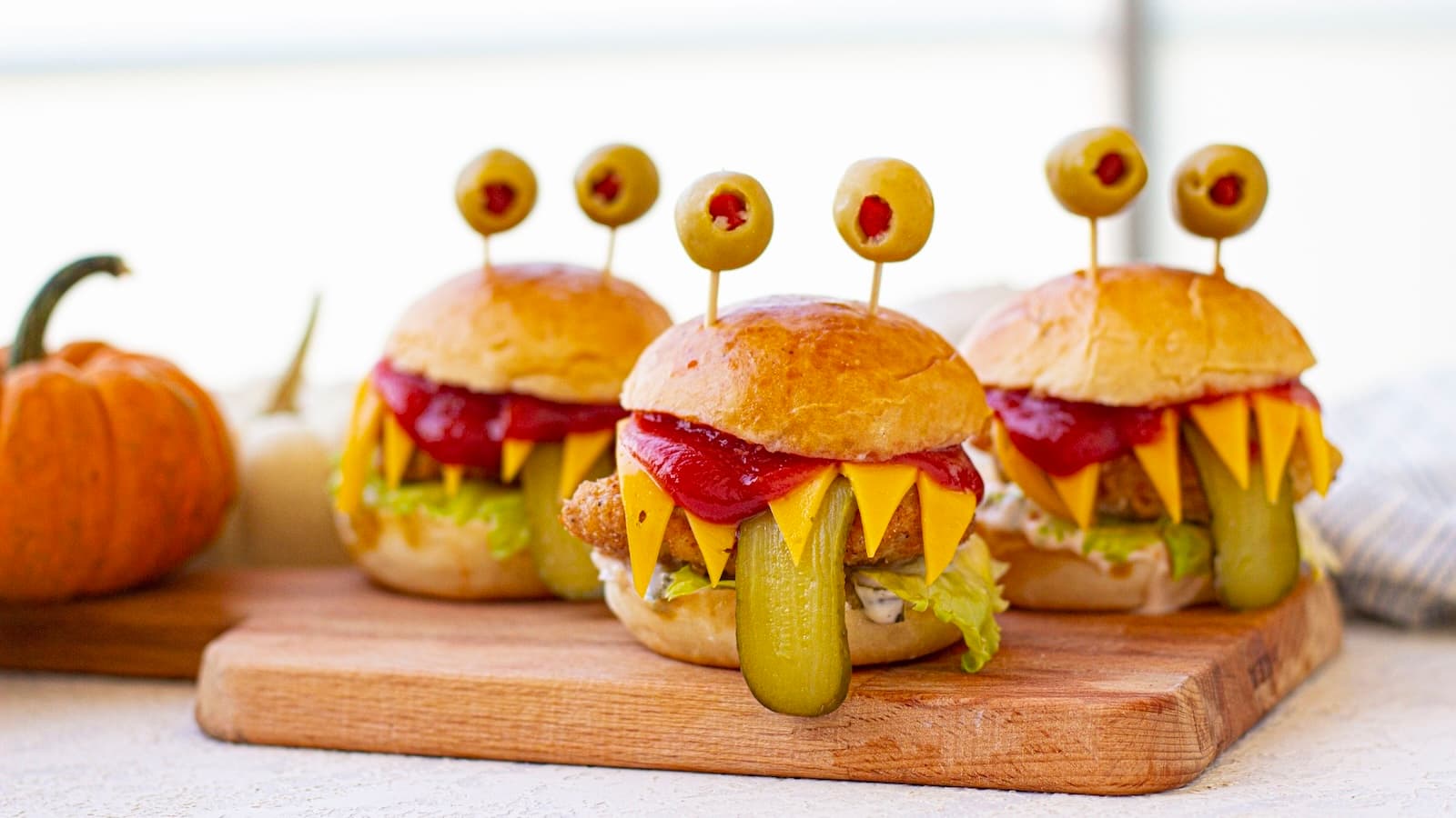 Three burgers with cheese, lettuce, pickles, and ketchup, designed to look like monster faces with olives on toothpicks as eyes, are placed on a wooden board.