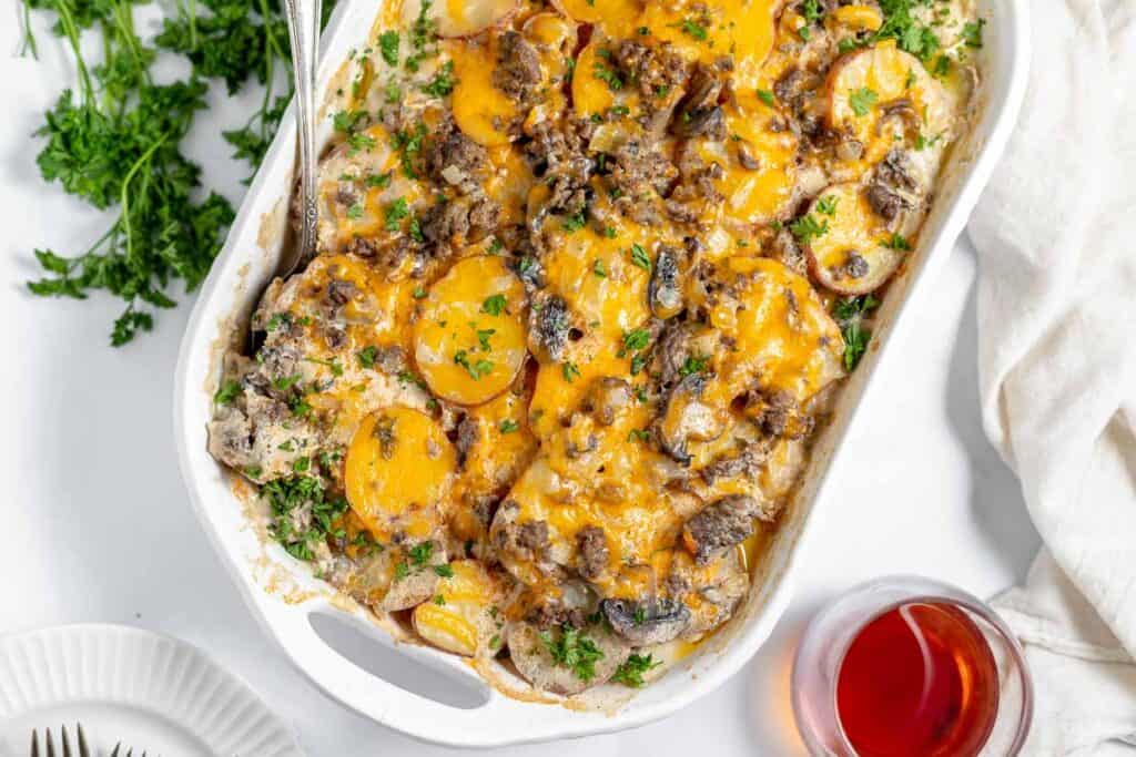 A casserole dish filled with baked vegetables, melted cheese, and sliced sausage, garnished with parsley. A glass of red beverage and some parsley are visible beside the dish.