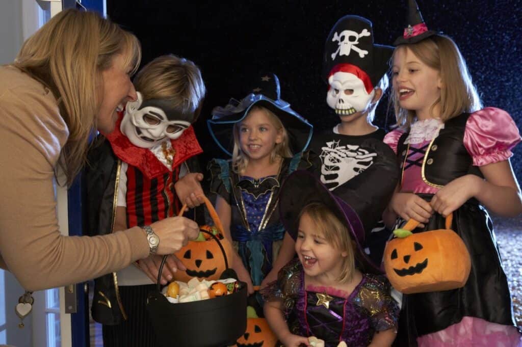 A woman distributes a mix of candy and non-candy trick-or-treat goodies to a group of children dressed in various Halloween costumes, each holding a pumpkin-shaped basket.