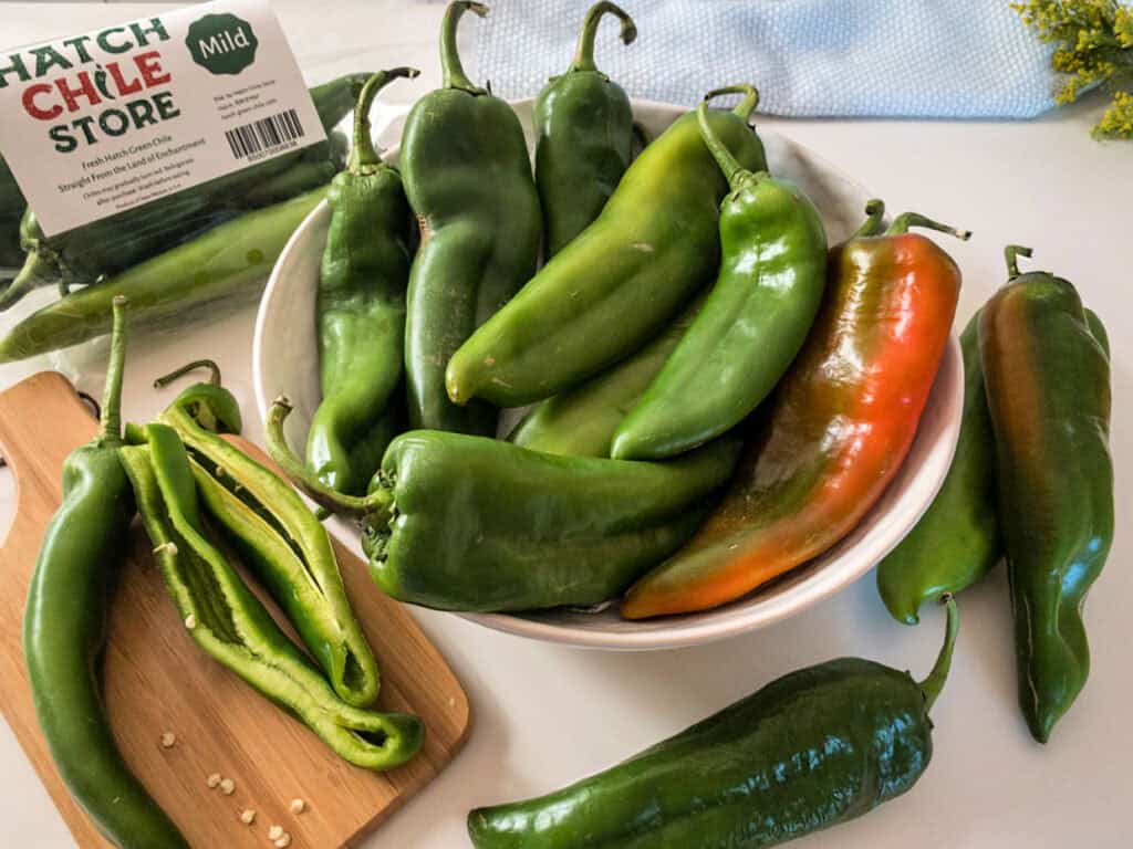 A bowl of fresh Hatch chile peppers on a white countertop, with a sliced chile on a cutting board next to it. A package labeled "Hatch Chile Store" is visible in the background.