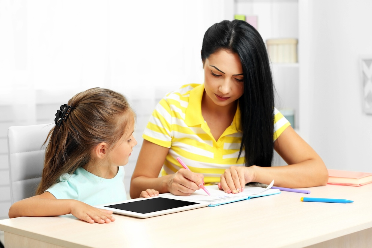 A woman is doing her girl's homework for her. The girl has a tablet, and the woman uses a pencil and a notebook. They are in a bright room with shelves and books in the background.