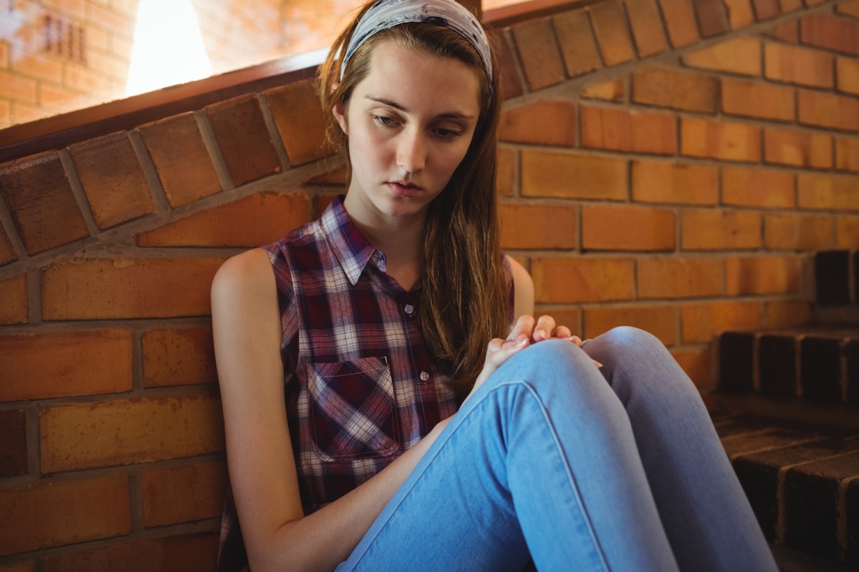 A woman in a plaid shirt and blue jeans sits on stairs against a brick wall, looking down pensively.