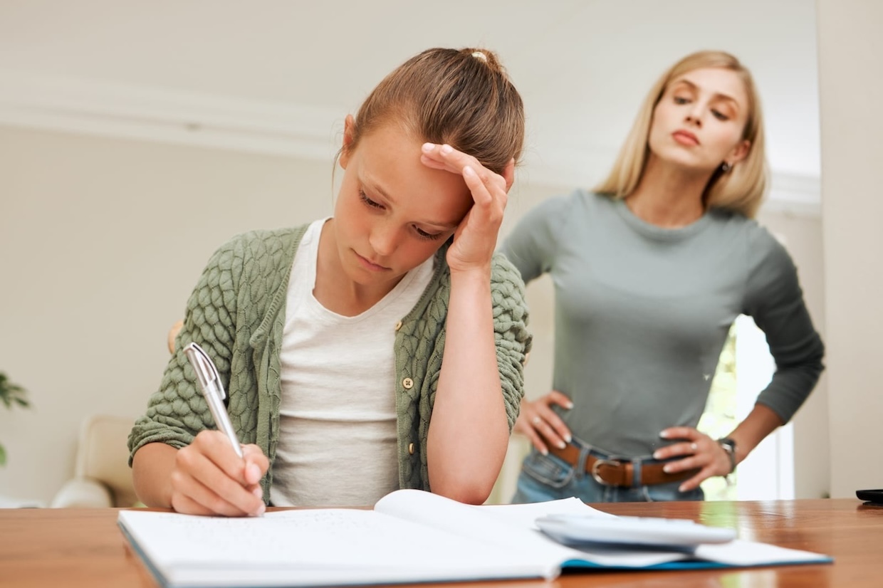 A young girl is doing homework at a table, looking frustrated with her hand on her forehead. A woman stands behind her with a stern expression and hands on her hips.