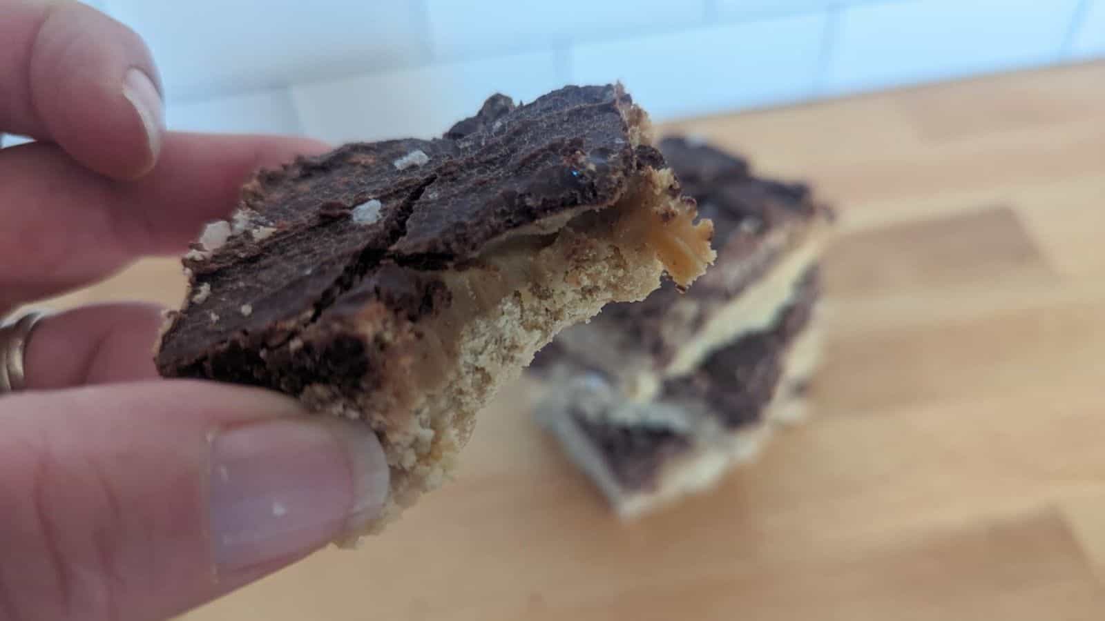 Image sjows a hand holds a partially bitten chocolate and caramel layered treat with a crumbly base, with more treats stacked in the background on a wooden surface.