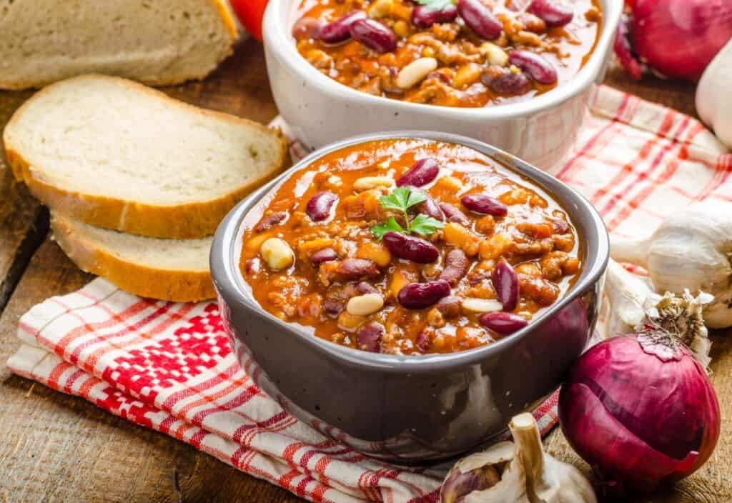 Two bowls of chili, garnished with herbs and beans, are placed next to sliced bread on a red and white checkered cloth, surrounded by garlic bulbs and red onions.