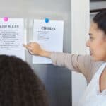 A woman points to a list of chores on a refrigerator door while talking to another person. A list of house rules is also posted next to the chores.