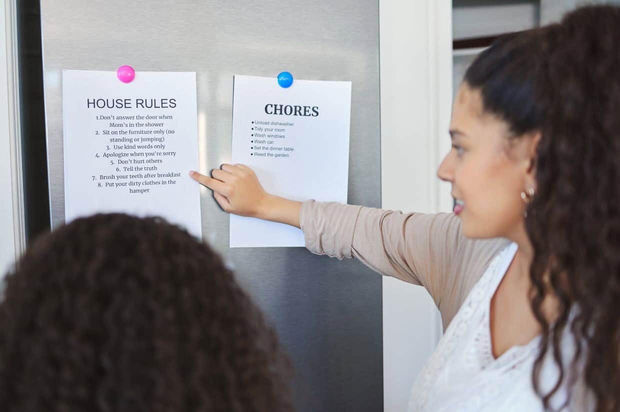 A woman points to a list of chores on a refrigerator door while talking to another person. A list of house rules is also posted next to the chores.