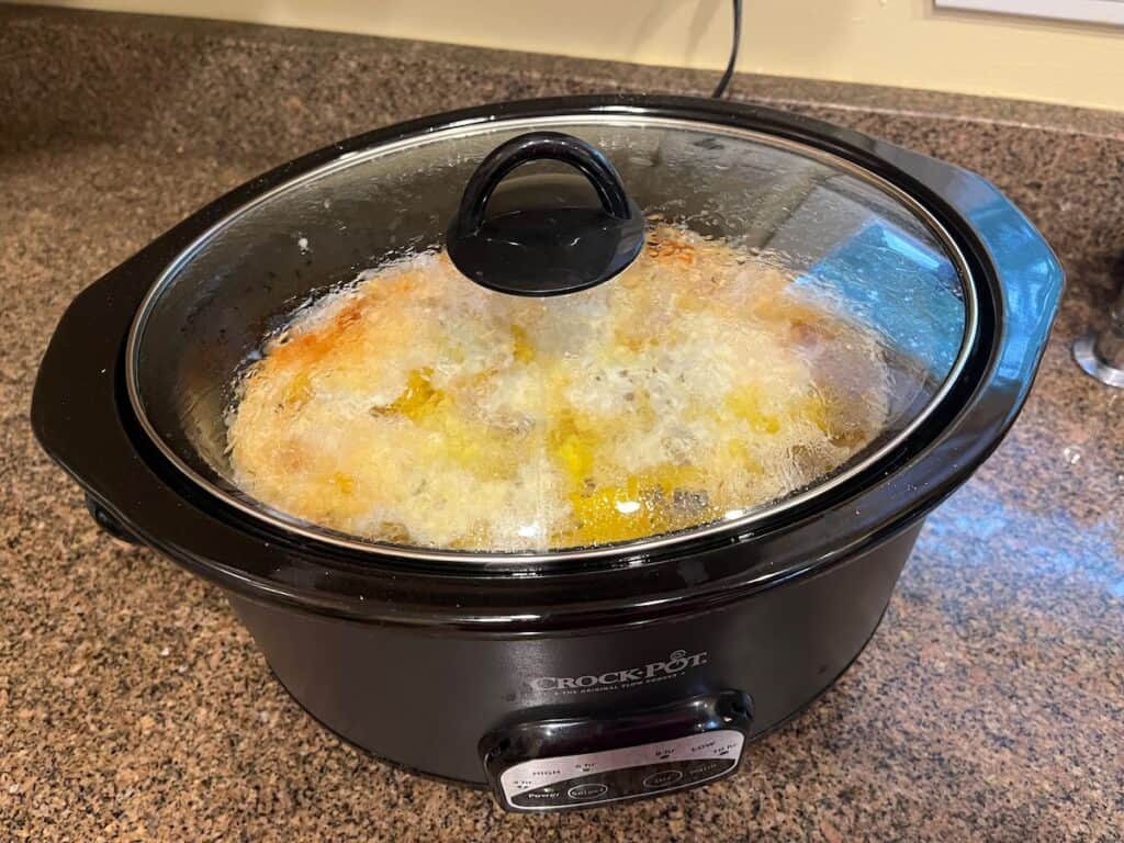 A black Crock-Pot slow cooker on a countertop is cooking a meal covered with a glass lid, showing condensation and bubbling liquid inside.