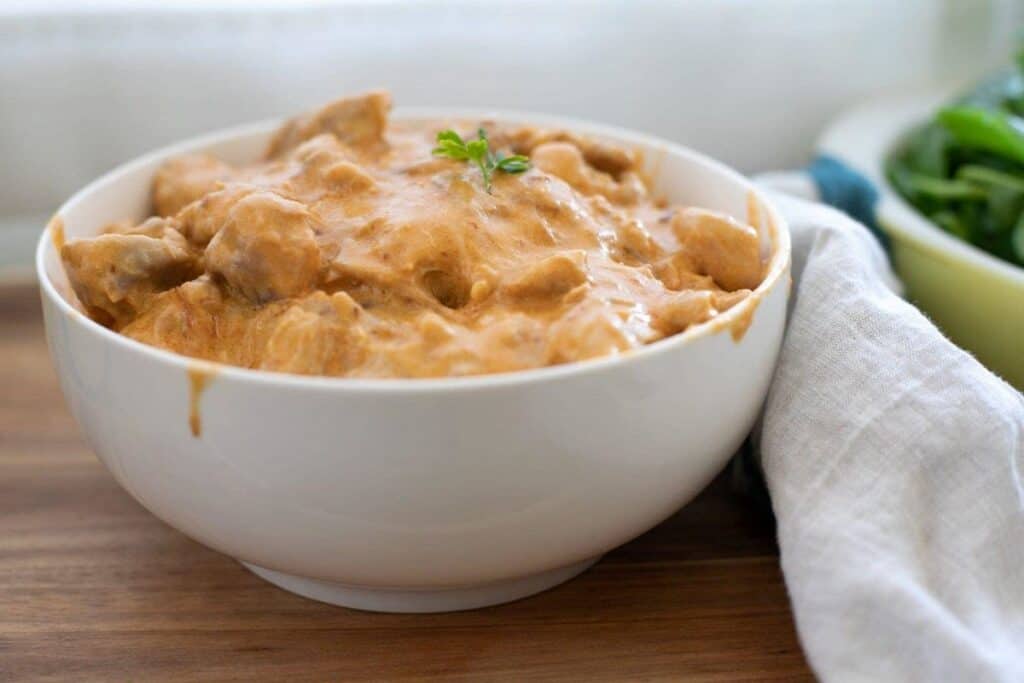 A white bowl filled with creamy butter chicken garnished with a small herb on top, placed on a wooden surface with a white napkin partially visible.