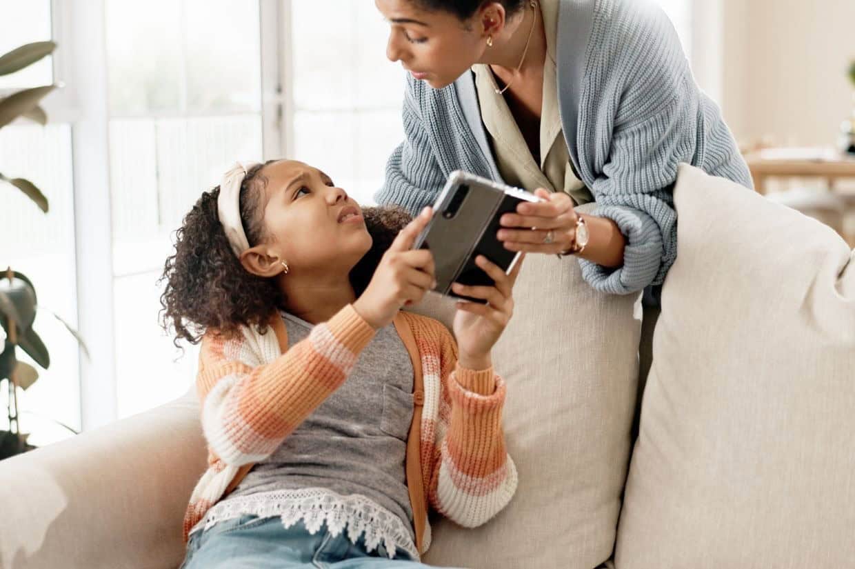 A woman in a cardigan is talking to a young girl sitting on a couch, who is holding a tablet.