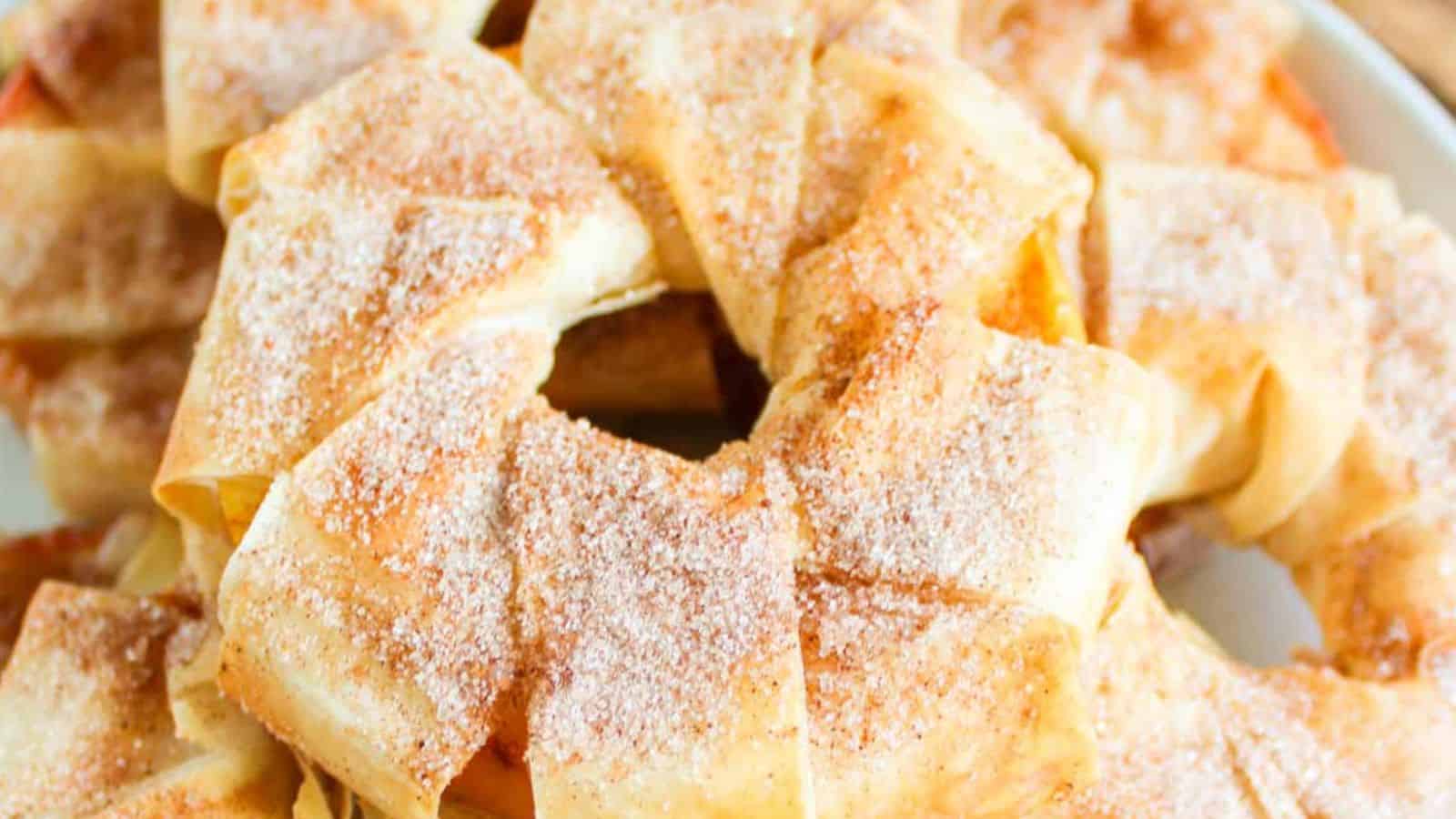 A close-up of a baked pastry ring dusted with powdered sugar, featuring multiple layers of golden-brown, flaky dough and hints of cinnamon apple strudel slices peeking through.