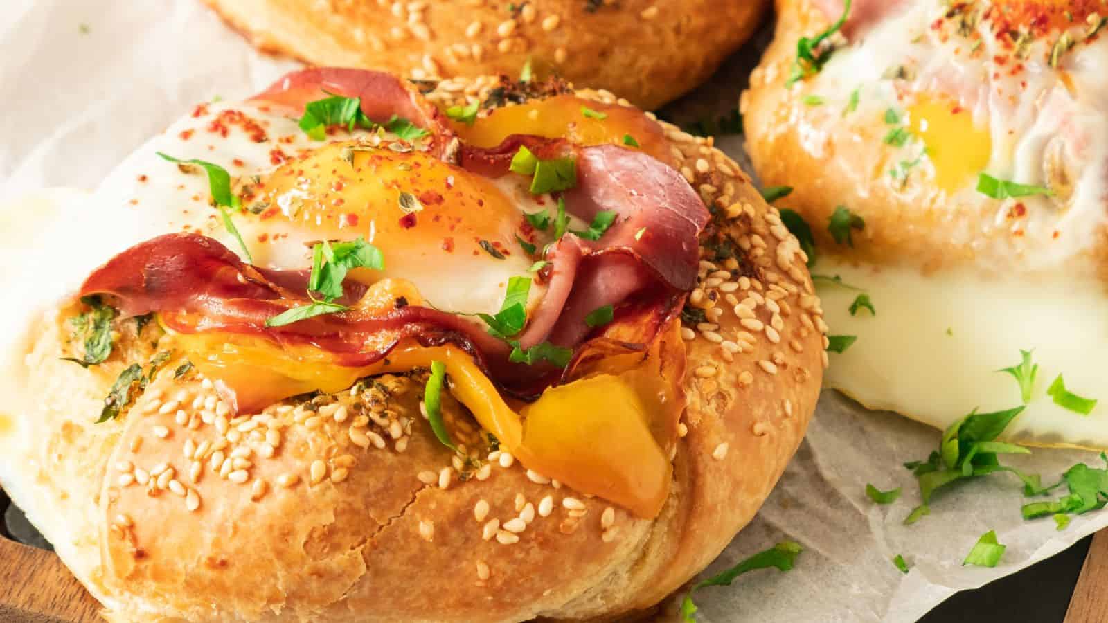 Close-up of a savory pastry topped with a sunny-side-up egg, sliced ham, yellow bell pepper, and garnished with chopped parsley, served on a sesame seed bun.