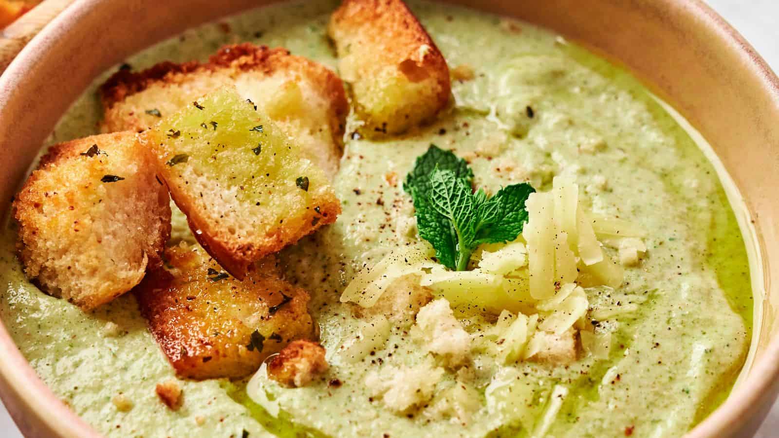 A close-up of a bowl of broccoli cheddar soup garnished with croutons, a sprig of mint, and grated cheese.