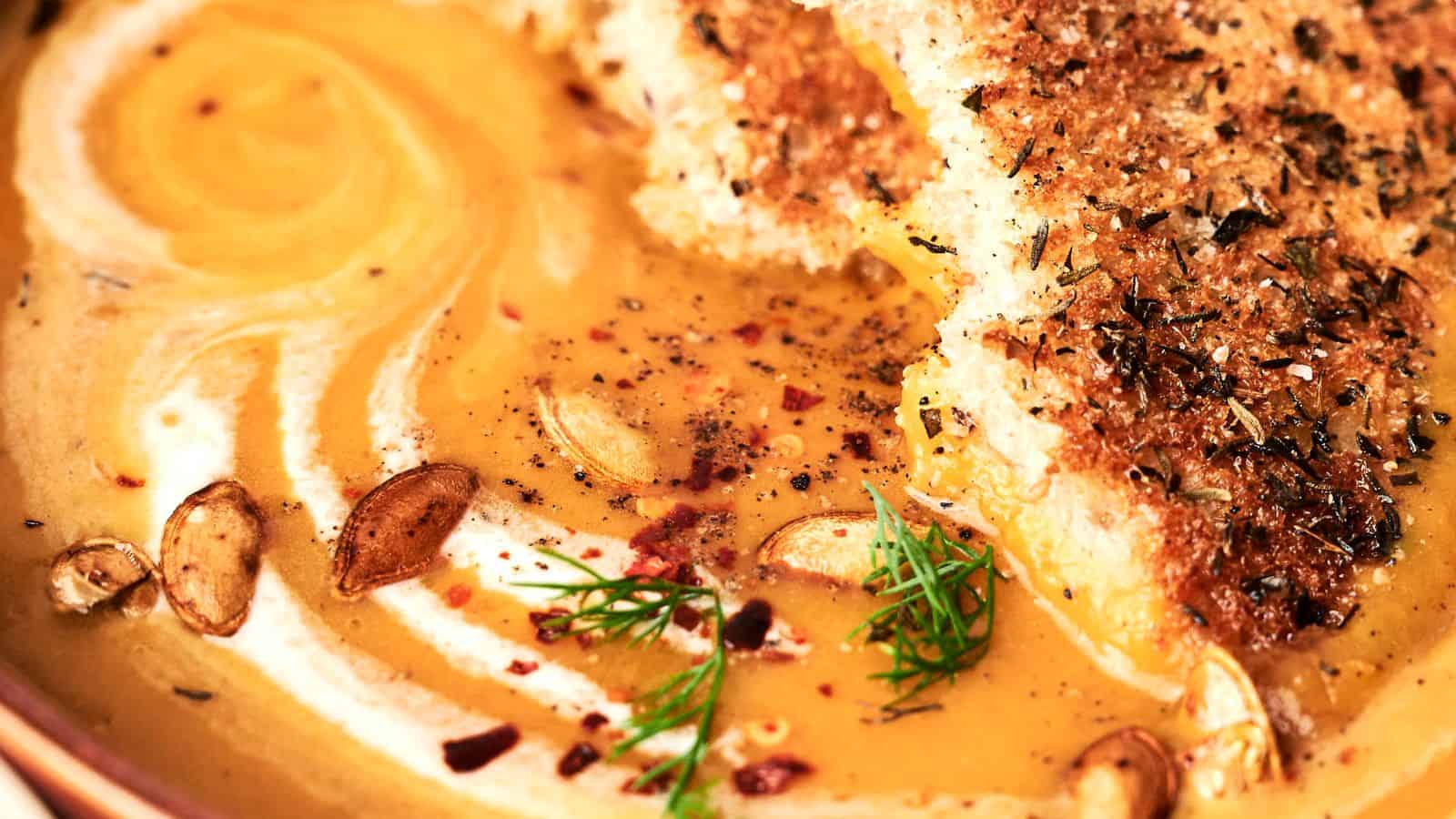 Close-up of a bowl of creamy butternut squash soup garnished with herbs, seeds, and two slices of seasoned bread. The soup has a swirled texture and a vibrant orange color.