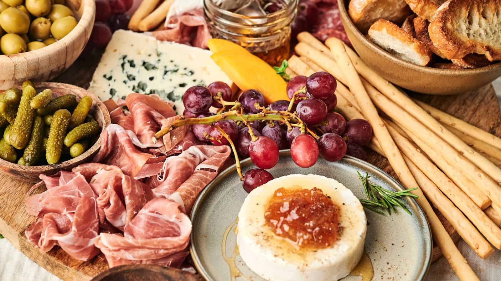 A charcuterie board featuring prosciutto, pickles, green olives, blue cheese, breadsticks, baguette slices, grapes, a jar of honey, and a round cheese topped with a dollop of jam.