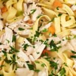 Close-up of a bowl of chicken noodle soup with shredded chicken, wide egg noodles, chopped vegetables, and garnished with parsley.