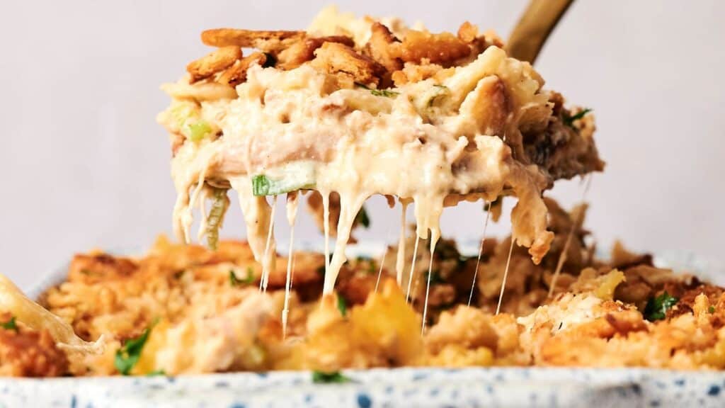 A spoon lifts a portion of a baked creamy casserole topped with crispy fried onions, with melted cheese stretching between the dish and the spoon.