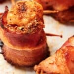 Close-up of several bacon-wrapped scallops secured with wooden toothpicks, resting on a white surface sprinkled with seasoning. The bacon is crispy, and the scallops have a browned, seasoned top.