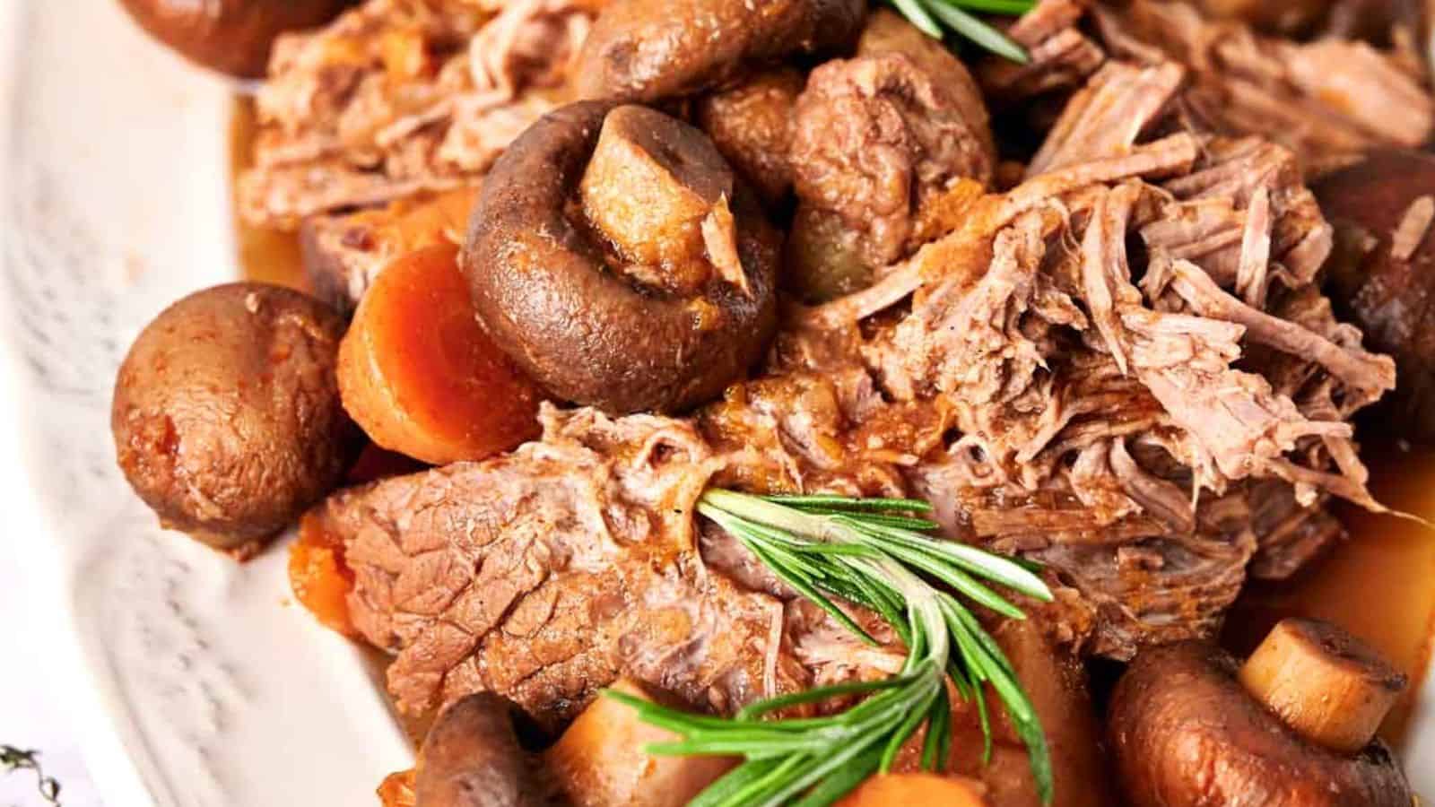 Close-up of a beef pot roast with mushrooms, carrots, and garnished with rosemary sprigs on a white plate.