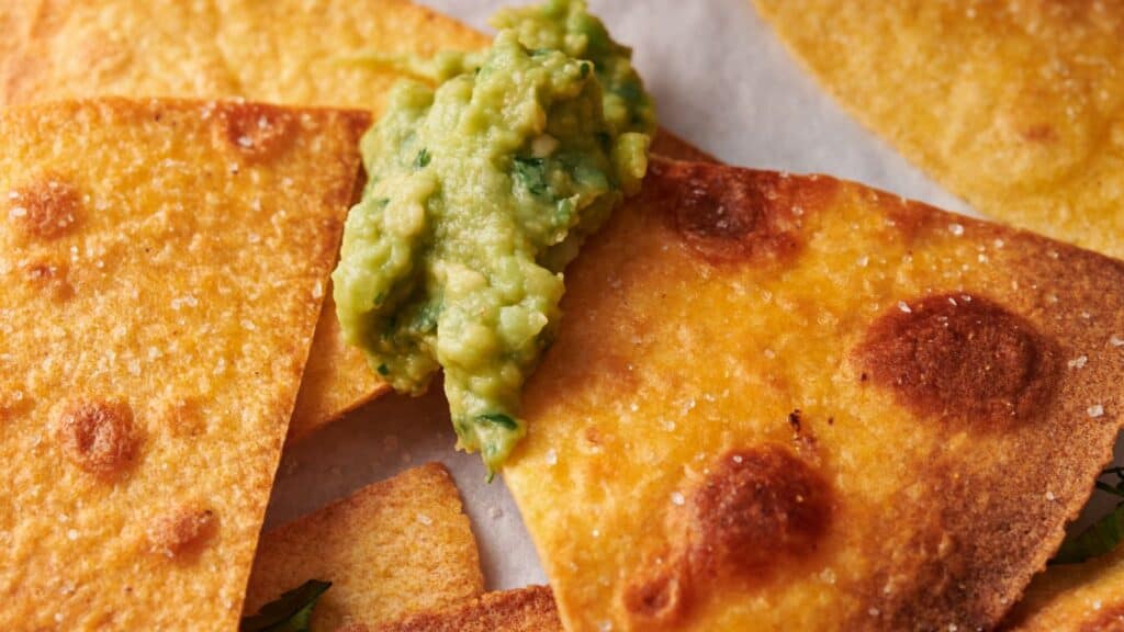 Close-up of golden-brown tortilla chips with a dollop of guacamole on top. The chips appear crispy with a few browned spots and a light sprinkle of salt.