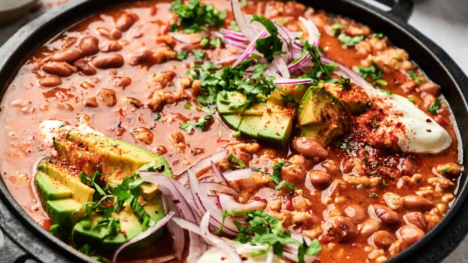 A close-up of a pot of chili with beans, avocado slices, red onion, sour cream, and garnished with chopped cilantro.