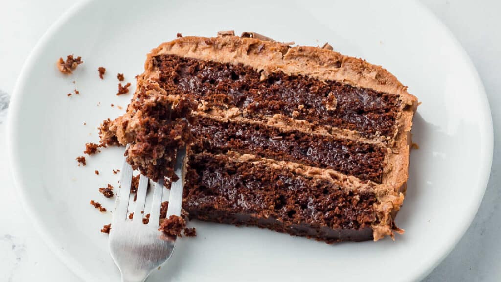 A partially eaten slice of chocolate layer cake with frosting is on a white plate. A fork with a bite of the cake is also on the plate.