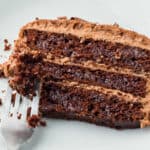 A partially eaten slice of chocolate layer cake with frosting is on a white plate. A fork with a bite of the cake is also on the plate.