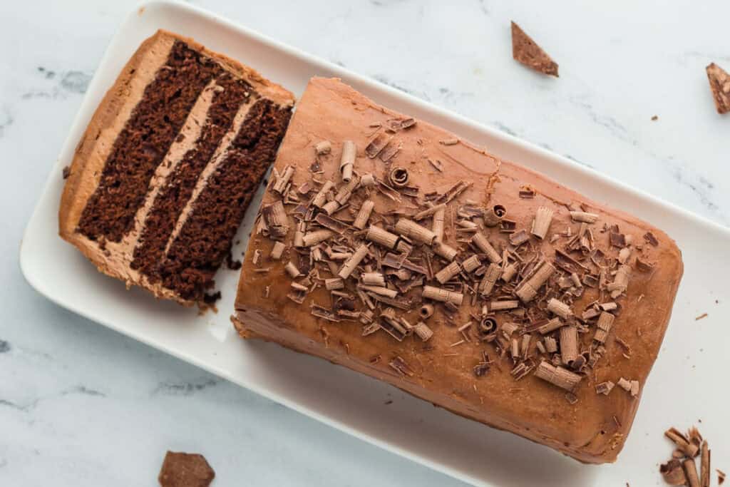 A sliced chocolate loaf cake with chocolate shavings on top, placed on a white rectangular platter. Some chocolate pieces are scattered around on the surface.