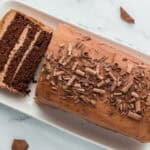 A sliced chocolate loaf cake with chocolate shavings on top, placed on a white rectangular platter. Some chocolate pieces are scattered around on the surface.