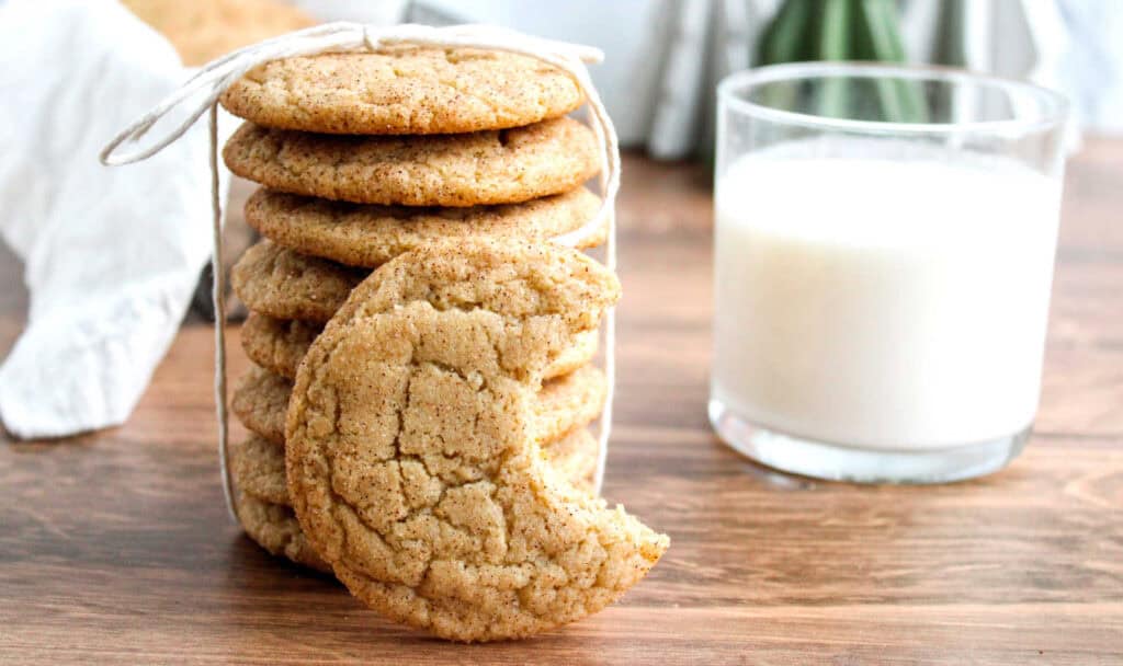 A stack of homemade cookies tied with a string, with one cookie showing a bite taken out of it, and a glass of milk on a wooden table.