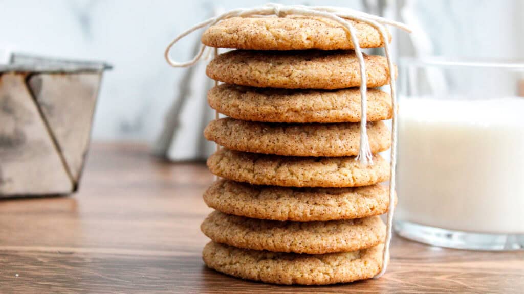 A stack of seven round cookies tied with a string, placed on a wooden surface with a glass of milk and a baking dish in the background.