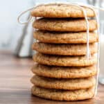 A stack of seven round cookies tied with a string, placed on a wooden surface with a glass of milk and a baking dish in the background.