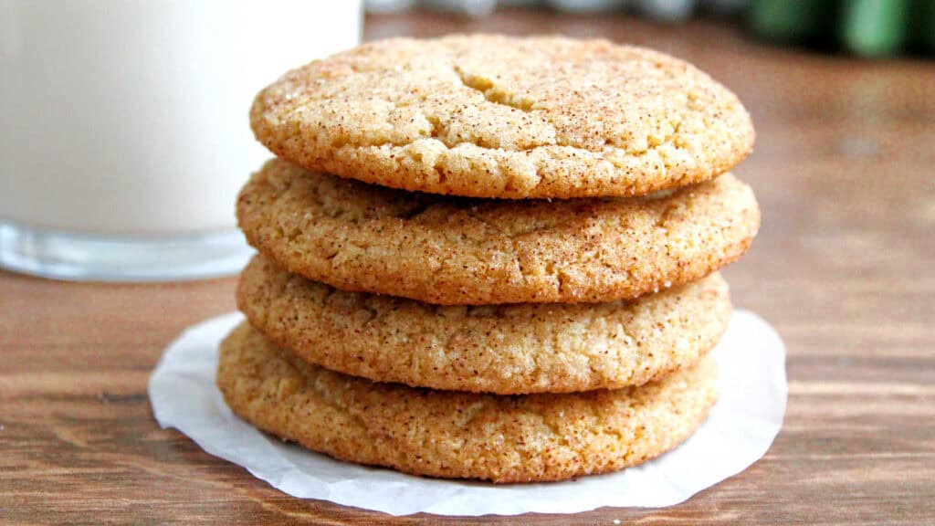 A stack of four snickerdoodle cookies on parchment paper with a glass of milk in the background.