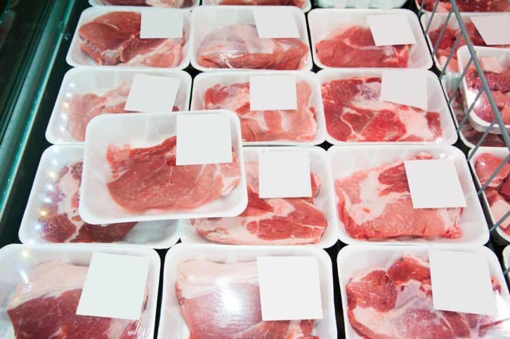 Pre-packaged cuts of raw meat are displayed in white trays with blank labels at a refrigerated grocery store section.