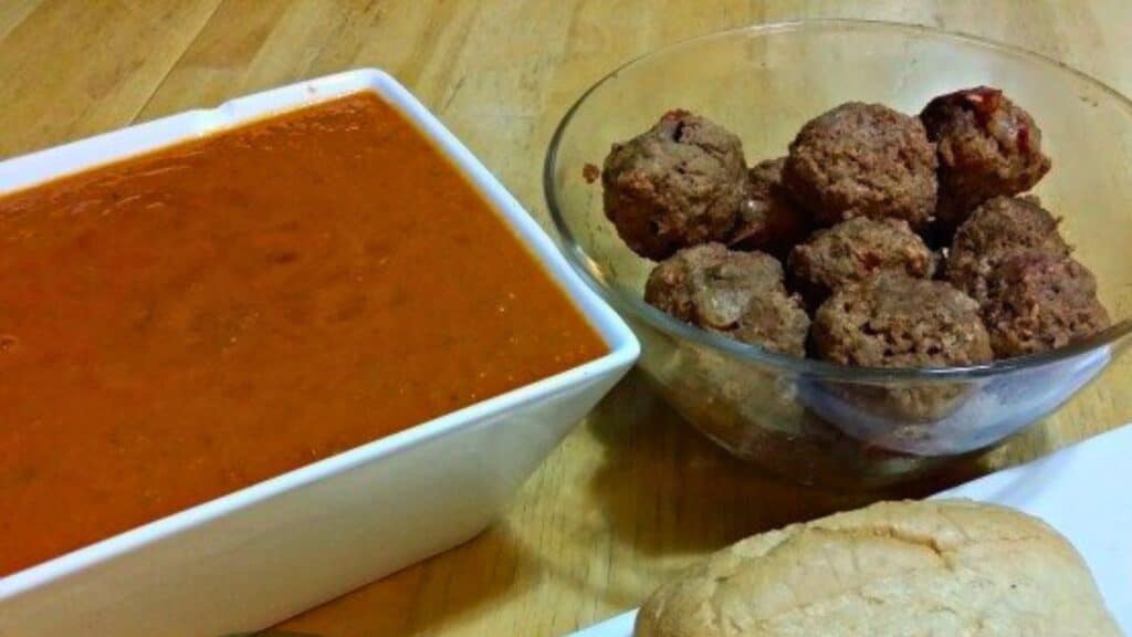 A rectangular white dish filled with tomato soup, a glass bowl with meatballs, and a bread roll on the right, all placed on a wooden surface.