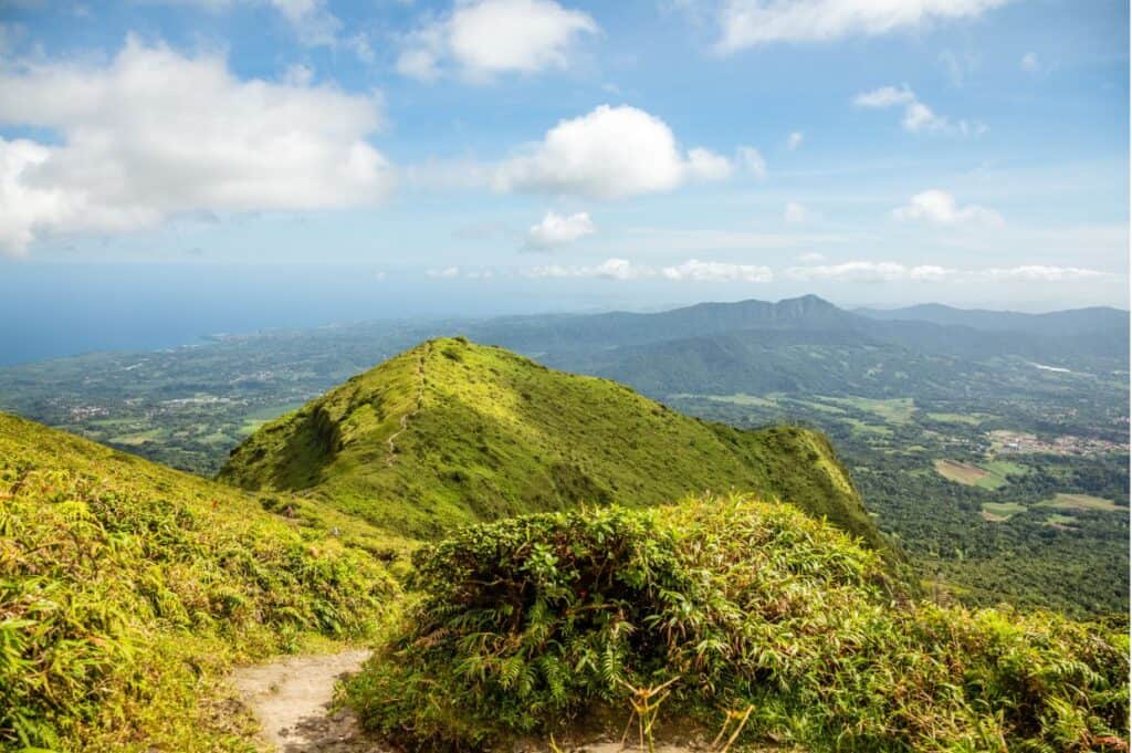 A scenic view of Martinique's lush landscapes with green mountainous terrain, distant hills, a coastline, and a partly cloudy sky.