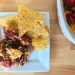 A plate with tortilla chips, black bean salsa, and shredded cheese on a wooden table next to a larger dish of the same dip garnished with diced tomatoes and green onions.