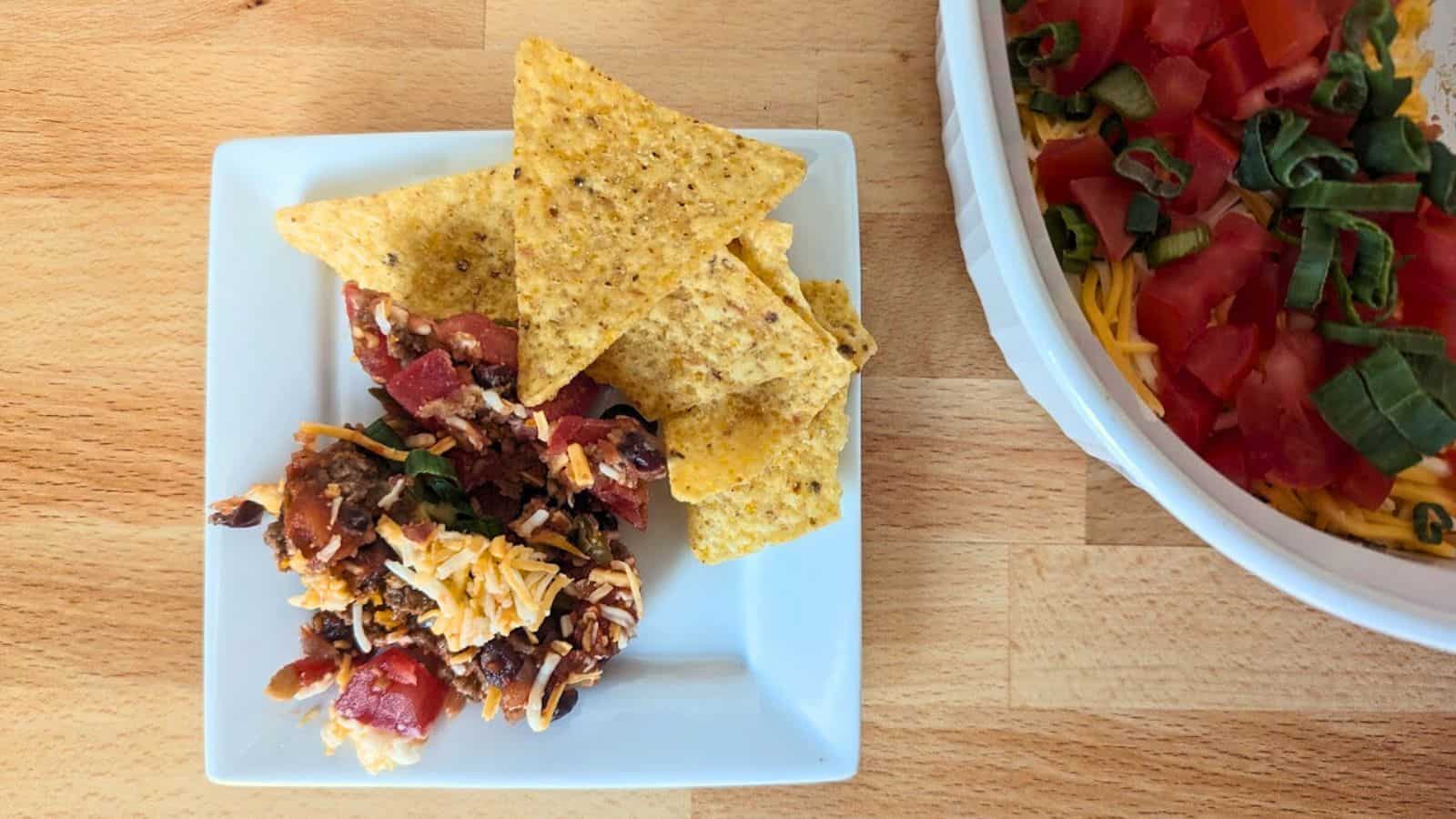 A plate with tortilla chips, black bean salsa, and shredded cheese on a wooden table next to a larger dish of the same dip garnished with diced tomatoes and green onions.