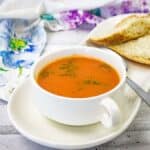 A white cup filled with tomato soup garnished with herbs sits on a matching saucer with a slice of bread.