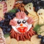 A Halloween-themed charcuterie board with meats, cheeses shaped like ghost faces, various fruits, nuts, and a cheese spread designed to look like a clown face.